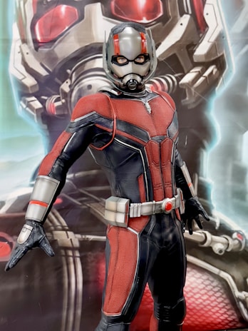 A person in a red and black superhero suit with a helmet, featuring silver accents, stands in a confident pose. The background shows an enlarged image of the helmet with red glowing eyes.