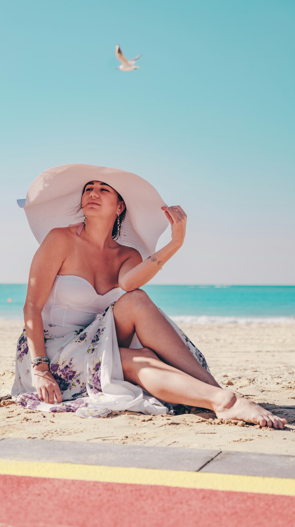 a woman sitting on the beach wearing a white hat