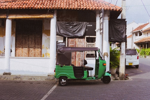 A green tuk-tuk is parked on a cobblestone street in front of an old building with white pillars and a red-tiled roof. The building has weathered wooden doors and is partially covered with black plastic sheets. There are signs with text on the wall and a potted plant to the side.