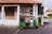 A green tuk-tuk is parked on a cobblestone street in front of an old building with white pillars and a red-tiled roof. The building has weathered wooden doors and is partially covered with black plastic sheets. There are signs with text on the wall and a potted plant to the side.