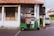 A green tuk-tuk is parked on a cobblestone street in front of an old building with white pillars and a red-tiled roof. The building has weathered wooden doors and is partially covered with black plastic sheets. There are signs with text on the wall and a potted plant to the side.
