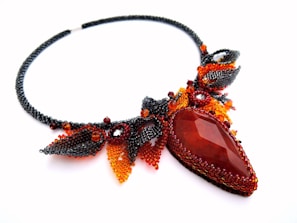 An intricately designed necklace featuring a large teardrop-shaped gemstone in a deep reddish hue. The necklace is adorned with beads in shades of black, red, and orange, arranged in a pattern that resembles leaves and abstract shapes. The craftsmanship is detailed and artistic, highlighting a blend of smooth and textured surfaces.