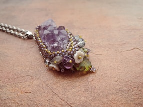 An intricately designed pendant featuring a cluster of purple crystals surrounded by small beads and floral decorations. The pendant is placed on a textured, earthy surface.