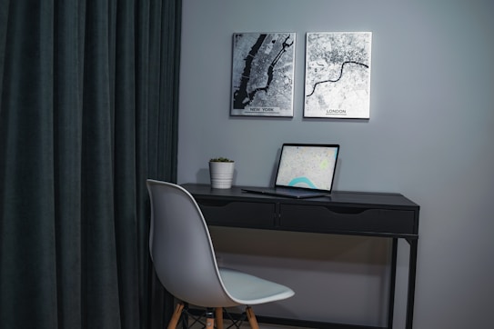 A modern workspace featuring a small, minimalist desk with a sleek, black finish. On the desk is an open laptop displaying a map screen. To the left, a white chair with wooden legs is tucked under the desk. A small potted plant adds a touch of greenery on the desk. Dark curtains serve as a backdrop on the left, while two black-and-white city map prints of New York and London are mounted on the wall above the desk.
