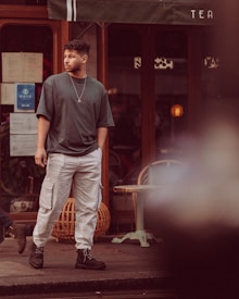 A person stands in front of a storefront, wearing a dark oversized t-shirt and light cargo pants with black boots. The background includes a window with visible papers and a partially obscured sign reading 'TEA'. There are plants and furniture visible through the window.