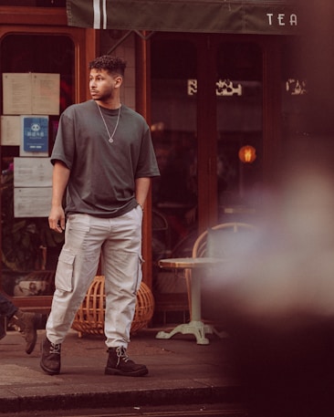 A person stands in front of a storefront, wearing a dark oversized t-shirt and light cargo pants with black boots. The background includes a window with visible papers and a partially obscured sign reading 'TEA'. There are plants and furniture visible through the window.