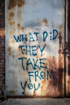 Graffiti with the message 'WHAT DID THEY TAKE FROM YOU' written in blue paint on a rusty, old metal door. The surface shows significant wear and rust, with patches of orange and brown. To the left is a section of a brick wall, and there's a padlock on the door to the right.