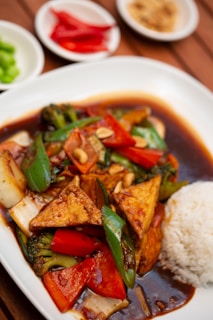 A plate of food featuring stir-fried vegetables with triangular pieces of tofu, served with a side of white rice. The dish is accompanied by a dark sauce and garnished with peanuts. In the background, there are small plates containing sliced red chilies, green peppers, and what appears to be ground peanuts or spices.