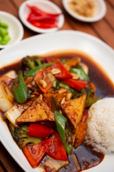 A plate of food featuring stir-fried vegetables with triangular pieces of tofu, served with a side of white rice. The dish is accompanied by a dark sauce and garnished with peanuts. In the background, there are small plates containing sliced red chilies, green peppers, and what appears to be ground peanuts or spices.
