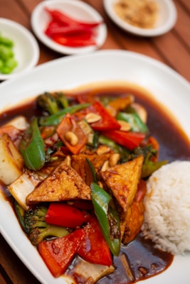 A plate of food featuring stir-fried vegetables with triangular pieces of tofu, served with a side of white rice. The dish is accompanied by a dark sauce and garnished with peanuts. In the background, there are small plates containing sliced red chilies, green peppers, and what appears to be ground peanuts or spices.