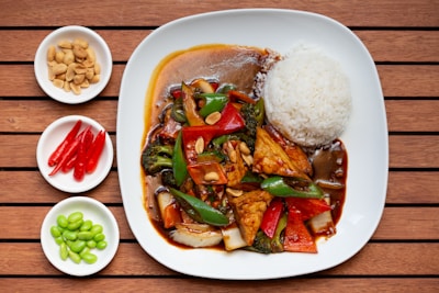 A plate of stir-fried vegetables and tofu with sauce is served alongside a mound of white rice. The dish is garnished with peanuts. Accompanying the main plate are three small bowls, each containing peanuts, red chili peppers, and edamame. The setting is on a wooden table, adding a warm and rustic feel to the presentation.