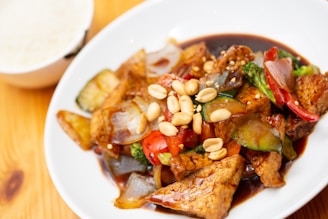 A delicious plate of stir-fried vegetables and tofu is garnished with peanuts and served on a white dish. The vibrant colors of red peppers, green broccoli, and glazed tofu are complemented by the sheen of a savory sauce. In the background, a bowl of rice is visible.