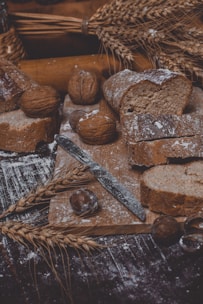 A rustic scene featuring sliced whole grain bread on a wooden board, surrounded by walnuts and chestnuts. A metal knife is placed on the board. There are stalks of wheat and flour scattered around, creating an artisanal, handcrafted atmosphere.