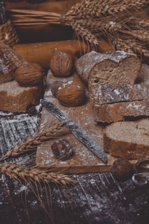 A rustic scene featuring sliced whole grain bread on a wooden board, surrounded by walnuts and chestnuts. A metal knife is placed on the board. There are stalks of wheat and flour scattered around, creating an artisanal, handcrafted atmosphere.