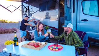 Three people are enjoying an outdoor gathering next to a recreational vehicle. Two of them are seated at a table with a cheerful spread of food including skewers, cheese, fruits, and wine, while another person is grilling in the background. The environment suggests a relaxed and sociable atmosphere.