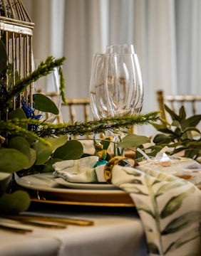 A beautifully set dining table with clear wine glasses, white plates with gold trim, and gold-colored cutlery. Decorative greenery, including leafy branches and sprigs, is elegantly arranged around the table. A cloth napkin with a green floral pattern is neatly placed, held together with a gold napkin ring.