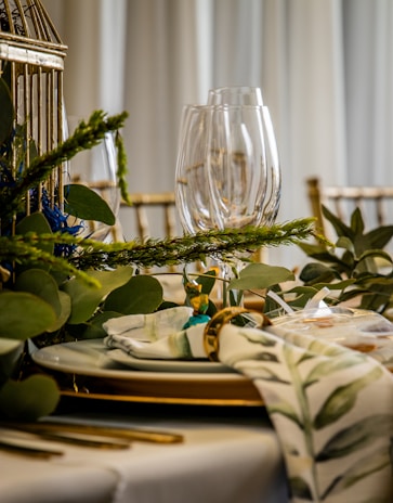 A beautifully set dining table with clear wine glasses, white plates with gold trim, and gold-colored cutlery. Decorative greenery, including leafy branches and sprigs, is elegantly arranged around the table. A cloth napkin with a green floral pattern is neatly placed, held together with a gold napkin ring.
