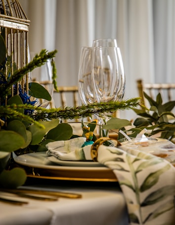 A beautifully set dining table with clear wine glasses, white plates with gold trim, and gold-colored cutlery. Decorative greenery, including leafy branches and sprigs, is elegantly arranged around the table. A cloth napkin with a green floral pattern is neatly placed, held together with a gold napkin ring.