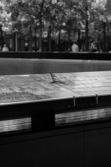 A close-up of a memorial plaque with names engraved on it, accompanied by a small American flag. The background shows trees and blurred figures, contributing to a solemn and reflective atmosphere.