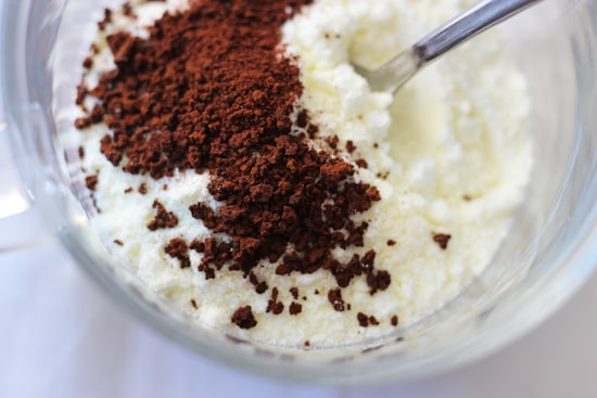 A bowl containing a mixture of white granular powder and dark brown coffee grounds with a metal spoon partially submerged.