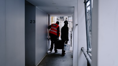 Two construction workers in safety vests and helmets are walking through a narrow passageway. One is carrying a paint bucket, and they appear to be engaged in work-related activities. The surrounding area is an urban setting with a mix of concrete and metal surfaces.