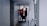 Two construction workers in safety vests and helmets are walking through a narrow passageway. One is carrying a paint bucket, and they appear to be engaged in work-related activities. The surrounding area is an urban setting with a mix of concrete and metal surfaces.