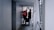Two construction workers in safety vests and helmets are walking through a narrow passageway. One is carrying a paint bucket, and they appear to be engaged in work-related activities. The surrounding area is an urban setting with a mix of concrete and metal surfaces.