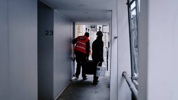 Two construction workers in safety vests and helmets are walking through a narrow passageway. One is carrying a paint bucket, and they appear to be engaged in work-related activities. The surrounding area is an urban setting with a mix of concrete and metal surfaces.
