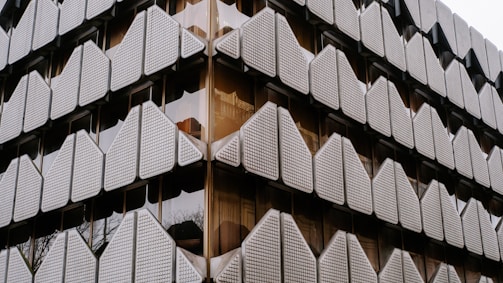 A building facade featuring a geometric, triangular pattern made of white, textured panels. The panels are arranged in a repetitive grid, reflecting a modern architectural style. The glass windows behind the panels add depth, providing a sleek and contemporary appearance.