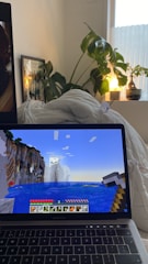 A cozy room with a laptop displaying a Minecraft game scene depicting a landscape with water and cliffs. The laptop is on a bed with white bedding. A large green plant sits beside the bed, and a small lamp emits a warm glow from a nearby window ledge. A framed picture is partially visible behind the plant.