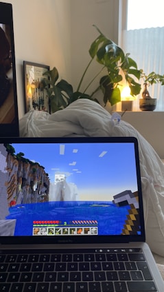 A cozy room with a laptop displaying a Minecraft game scene depicting a landscape with water and cliffs. The laptop is on a bed with white bedding. A large green plant sits beside the bed, and a small lamp emits a warm glow from a nearby window ledge. A framed picture is partially visible behind the plant.