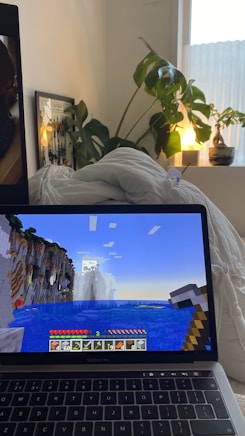 A cozy room with a laptop displaying a Minecraft game scene depicting a landscape with water and cliffs. The laptop is on a bed with white bedding. A large green plant sits beside the bed, and a small lamp emits a warm glow from a nearby window ledge. A framed picture is partially visible behind the plant.