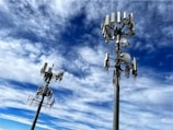 Two tall communication towers with multiple antennas and satellite dishes are positioned against a backdrop of a vibrant blue sky filled with scattered white clouds.