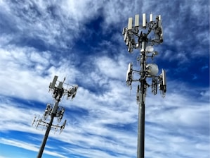 Two tall communication towers with multiple antennas and satellite dishes are positioned against a backdrop of a vibrant blue sky filled with scattered white clouds.