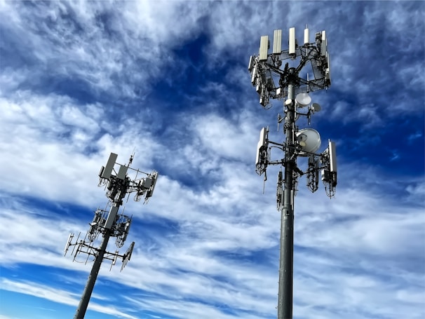 Two tall communication towers with multiple antennas and satellite dishes are positioned against a backdrop of a vibrant blue sky filled with scattered white clouds.