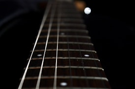 The image features a close-up of a guitar neck with visible strings and frets. The perspective is angled, focusing on the length of the neck, converging in the background. A blurred background enhances the sharpness of the strings and frets.
