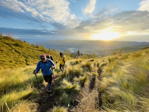 Two hikers in colorful outdoor gear walk along a grassy trail on a hillside, with trekking poles in hand. The landscape is bathed in the golden light of a sunrise or sunset, casting a warm glow over the scene. The horizon reveals rolling hills under a sky filled with dramatic clouds.
