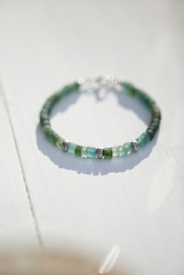 A beaded bracelet with various shades of green and blue beads interspersed with small silver accents is lying on a light-colored surface. The lighting creates soft shadows, giving a gentle and serene appearance.