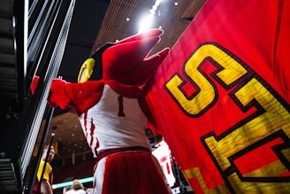 A mascot in a red and white costume stands holding a large red flag with yellow lettering. The setting appears to be an indoor sports arena, with a ceiling featuring spotlights. People can be seen in the background, possibly spectators or event staff.