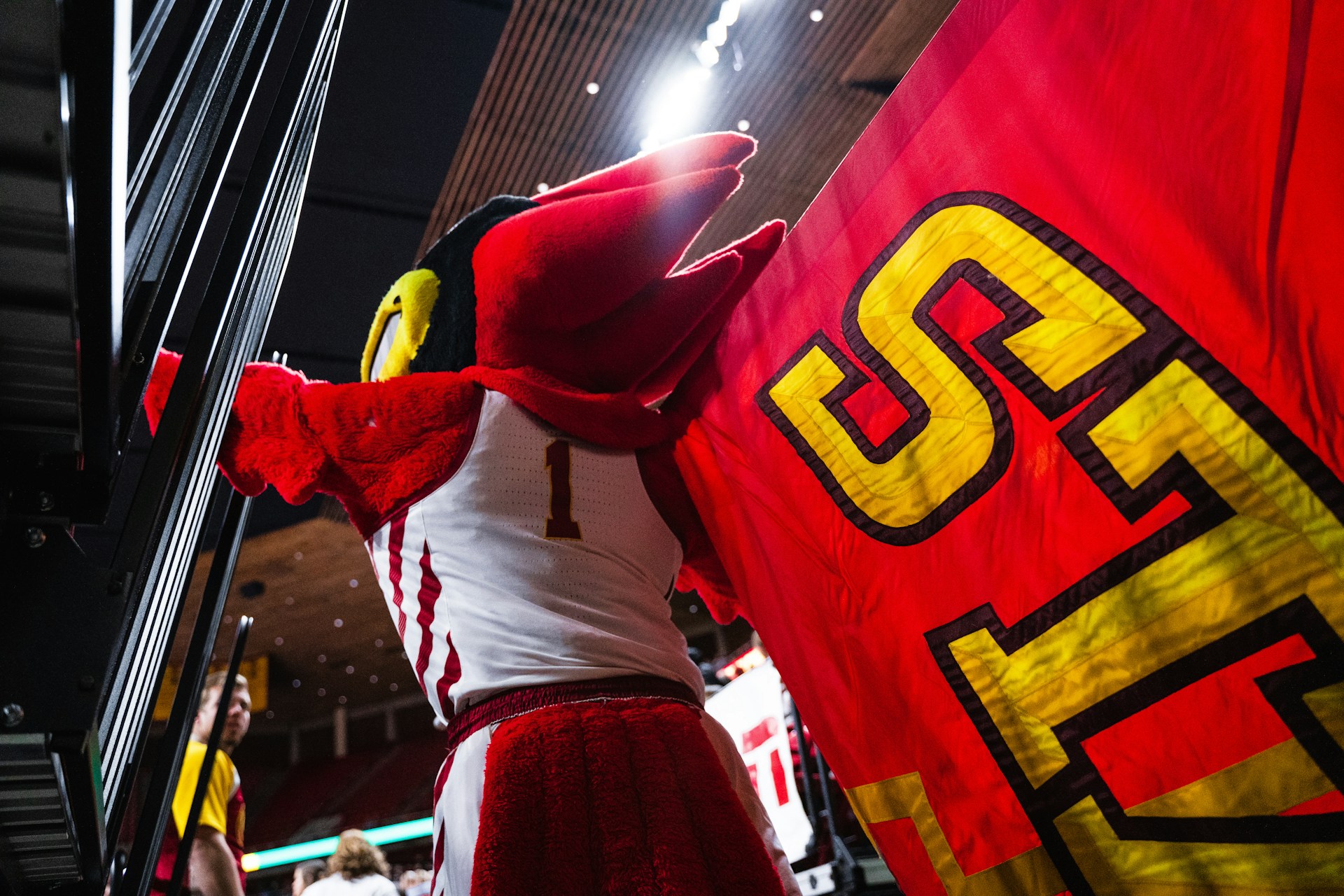 A side-splitting scene of a mascot tripping over its oversized feet during a halftime show, crowd laughing in the background.