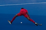 A field hockey player wearing a red uniform is stretching wide to reach a ball on a blue turf field. The player is bent over with one leg extended forward and the other backward, preparing to play the ball with a hockey stick.