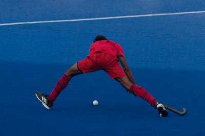 A field hockey player wearing a red uniform is stretching wide to reach a ball on a blue turf field. The player is bent over with one leg extended forward and the other backward, preparing to play the ball with a hockey stick.
