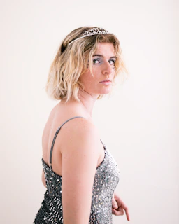 A person with short blonde hair wearing a sparkling, sequined dress and a tiara. They are posed in a side profile, looking towards the camera with a neutral expression.