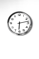 A simple wall clock with a white face and black numbers, featuring a minimalist design. It has a metal rim and traditional clock hands indicating the time. The background is plain and white, enhancing the clock's clean and classic appearance.