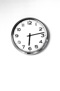 A simple wall clock with a white face and black numbers, featuring a minimalist design. It has a metal rim and traditional clock hands indicating the time. The background is plain and white, enhancing the clock's clean and classic appearance.