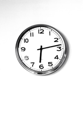 A simple wall clock with a white face and black numbers, featuring a minimalist design. It has a metal rim and traditional clock hands indicating the time. The background is plain and white, enhancing the clock's clean and classic appearance.