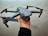 A gray drone with four propellers is being held in one hand against a light textured background. The drone has orange accents on the propeller tips and features a camera mounted on the front.