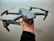 A gray drone with four propellers is being held in one hand against a light textured background. The drone has orange accents on the propeller tips and features a camera mounted on the front.