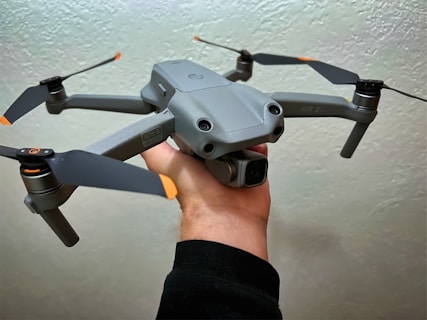 A gray drone with four propellers is being held in one hand against a light textured background. The drone has orange accents on the propeller tips and features a camera mounted on the front.