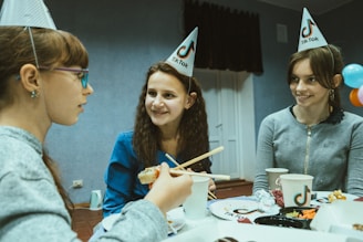 Three young girls are sitting around a table, wearing TikTok-themed party hats. They are engaging in a lively conversation and appear to be enjoying some food and drinks, with one girl using chopsticks to eat sushi.